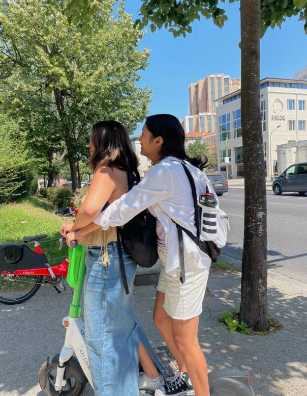 Two people in backpacks ride a white and green scooter on a sunny day.