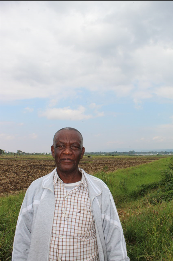 A man looks at the camera and stands in front of a farm field surrounded by tall grass.