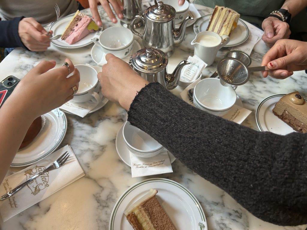 Hands reach out towards a circle of teacups around a silver teapot. There are also plates with cake on them.