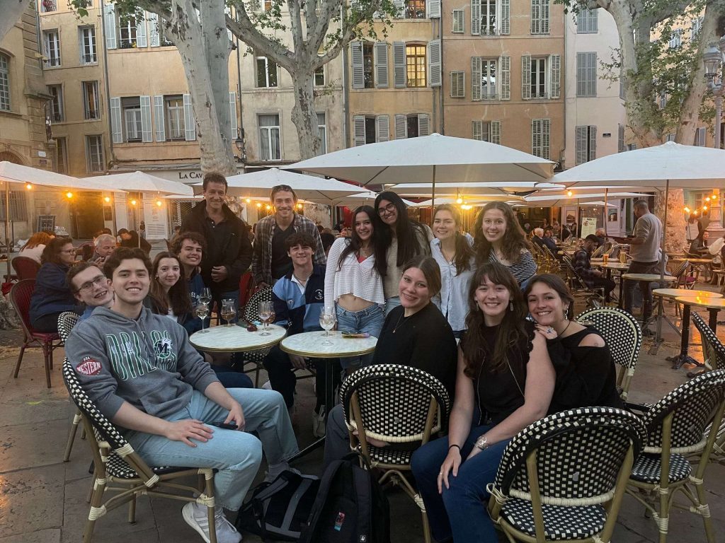 About a dozen individuals gathered at an outdoor eating space. They are sitting and standing aroud several small, circular tables.