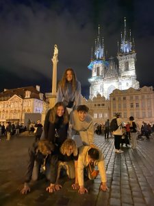 Six individuals posing in a human pyramid in a large square at night. A birhgtly lit church is behind them.