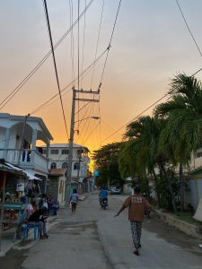 A main street in La Ciénaga, Dominican Republic. It is sunset and there are buildings on the left side of the street and people walking down the road.