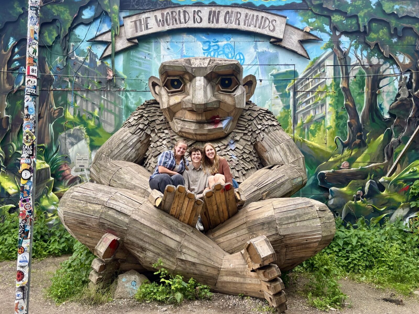 Three people smile and pose on a giant wooden statue of a troll sitting cross-legged in bushes.