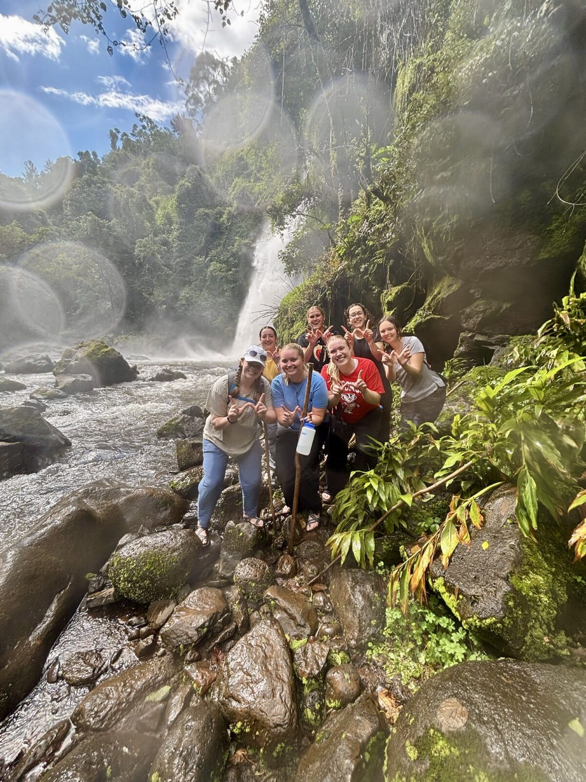 Seven students standing on wet rocks by lush greenery in front of a waterfall.