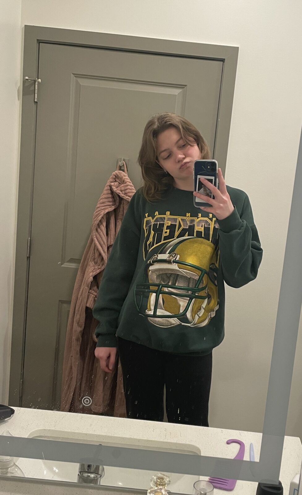 A student smiles at the camera in a mirror selfie. They are wearing a green Green Bay Packers sweatshirt and have their head tilted to the side