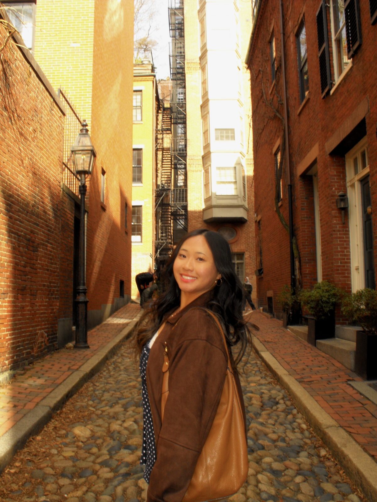 A student smiles at the camera. She has long, dark hair and has a light brown bag on her shoulder.