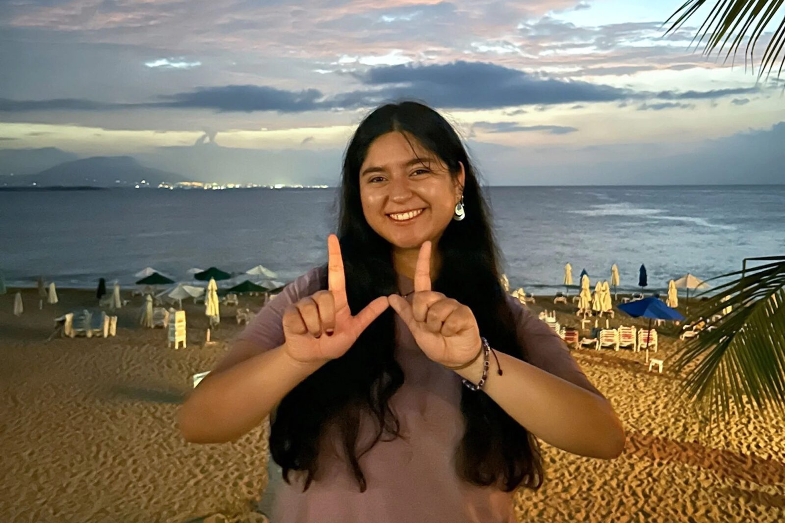 A student with long dark hair standing on a beach and makes a "W" with her fingers. The sun is setting behind her.
