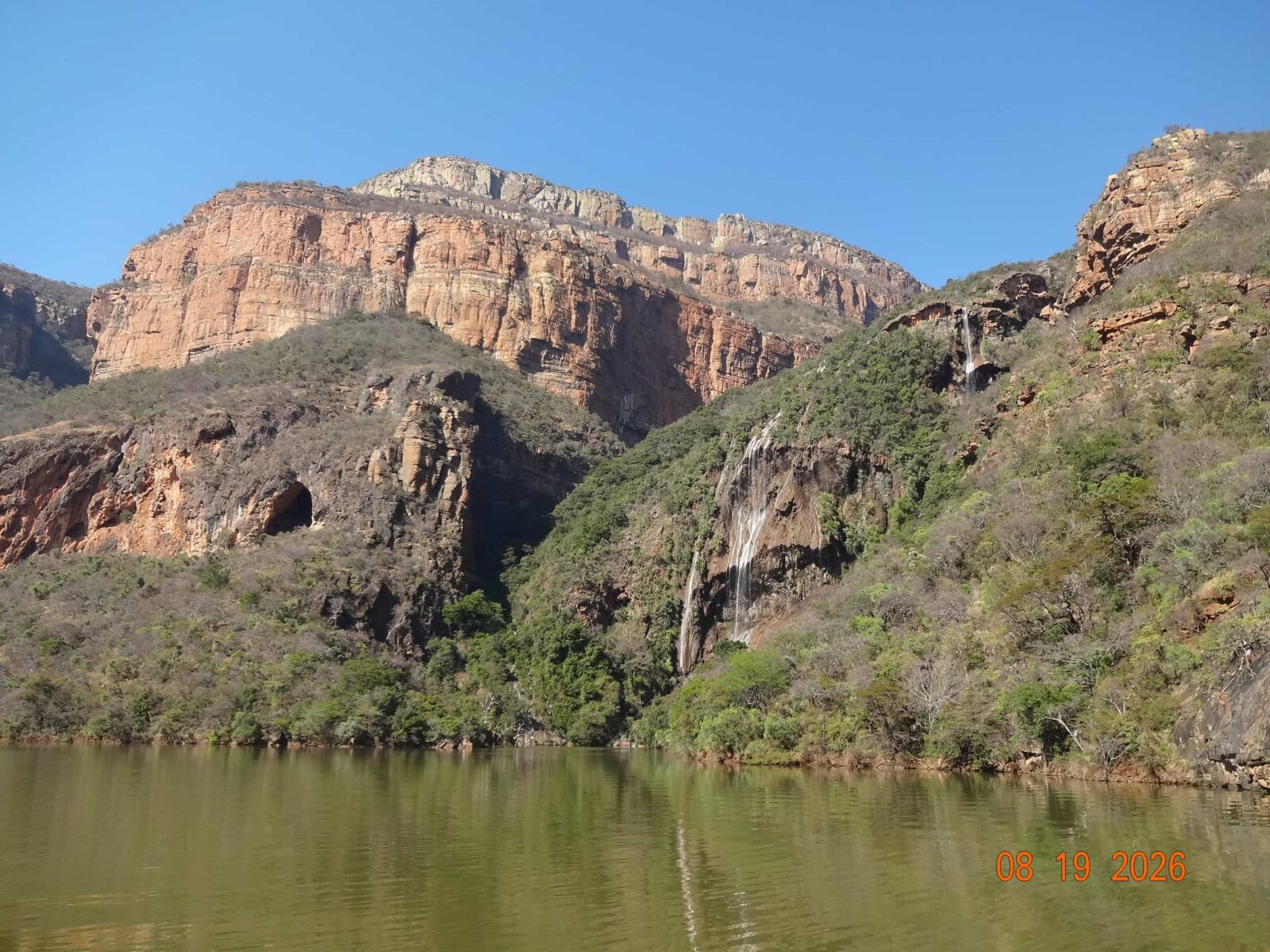 Waterfalls running over steep, shrub-covered hills into a larger body of water, with tall, rocky cliffs behind.