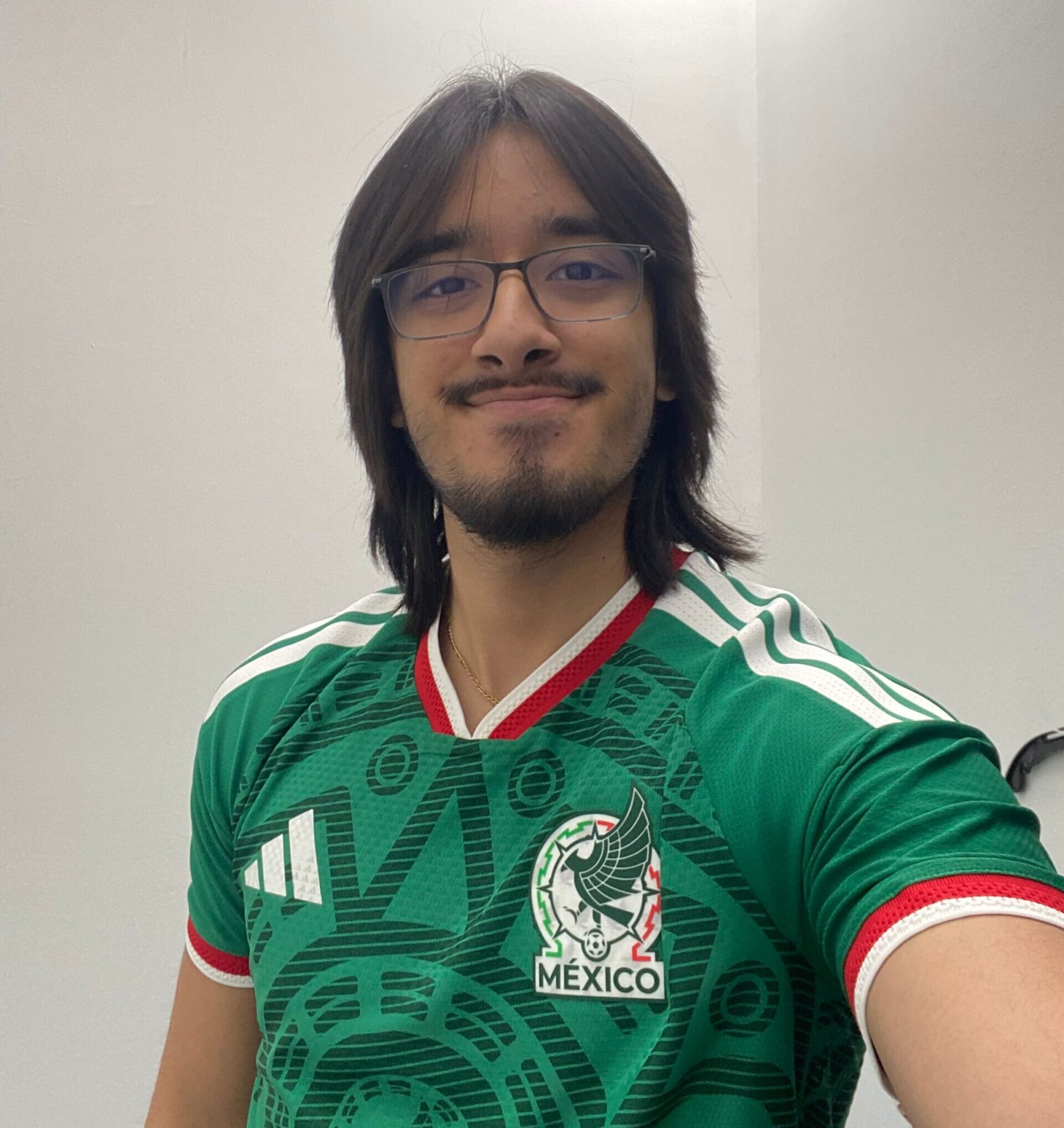 A man poses for a selfie photo in front of a white wall, wearing a green Mexico soccer jersey.