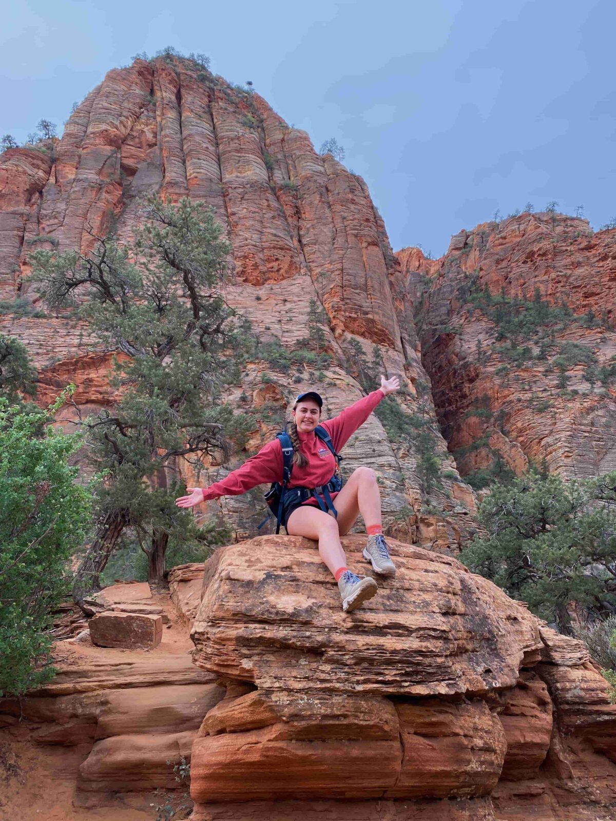 A woman in a red shirt sits on top of a large rock in a desert cave smiling at the camera.