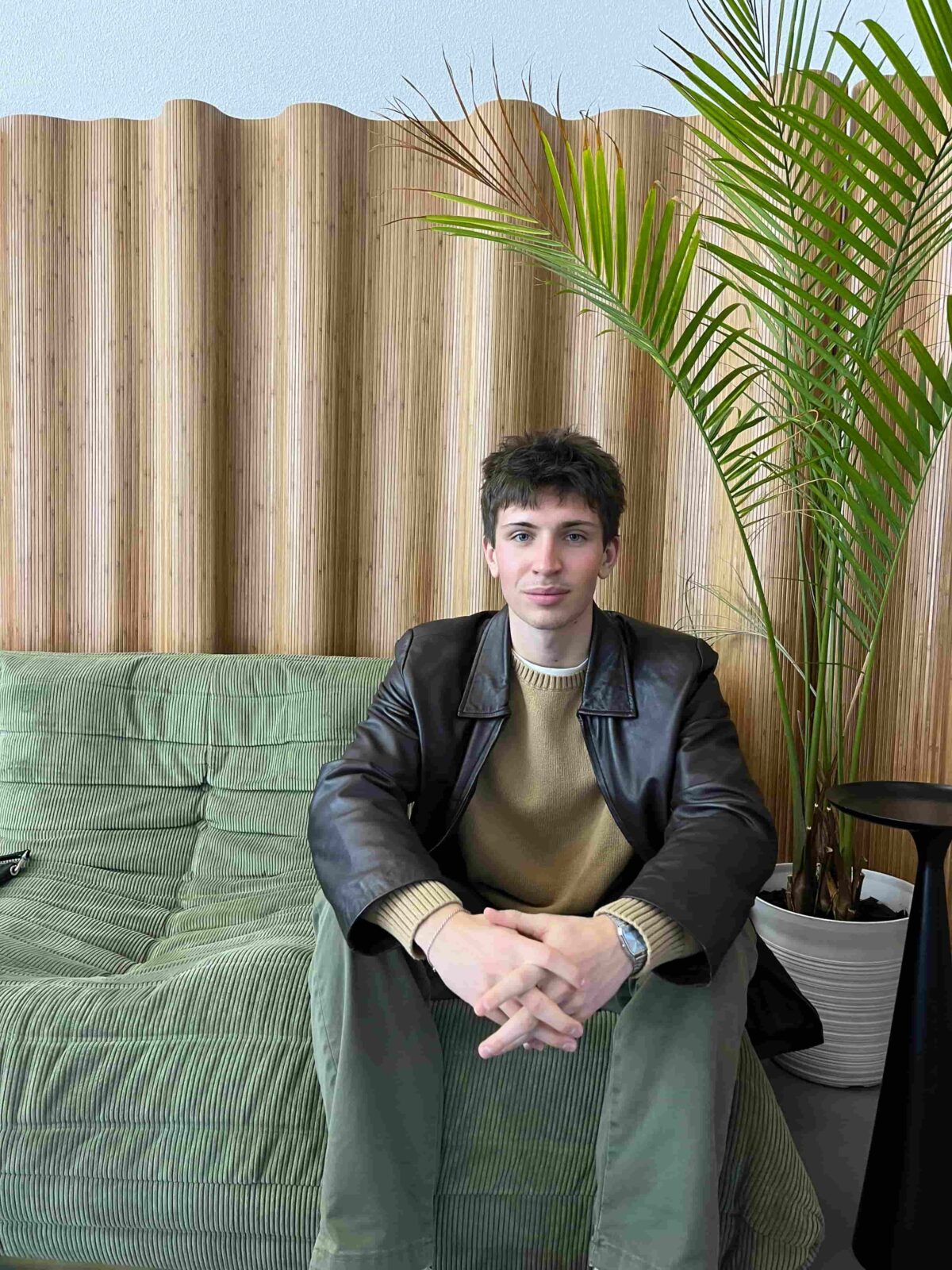 A person sits on a couch wearing a leather jacket and tam sweater with a plant to their left.