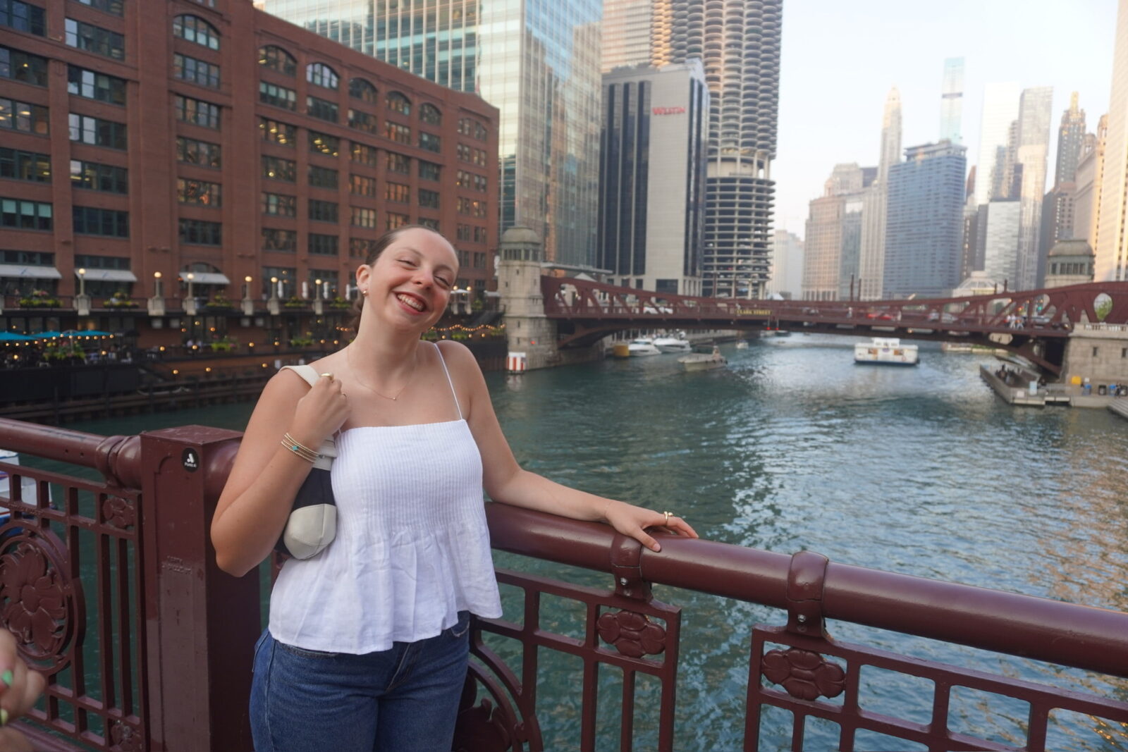 A woman wearing a white shirt and jeans smiles at the camera while standing on a bridge over a river.