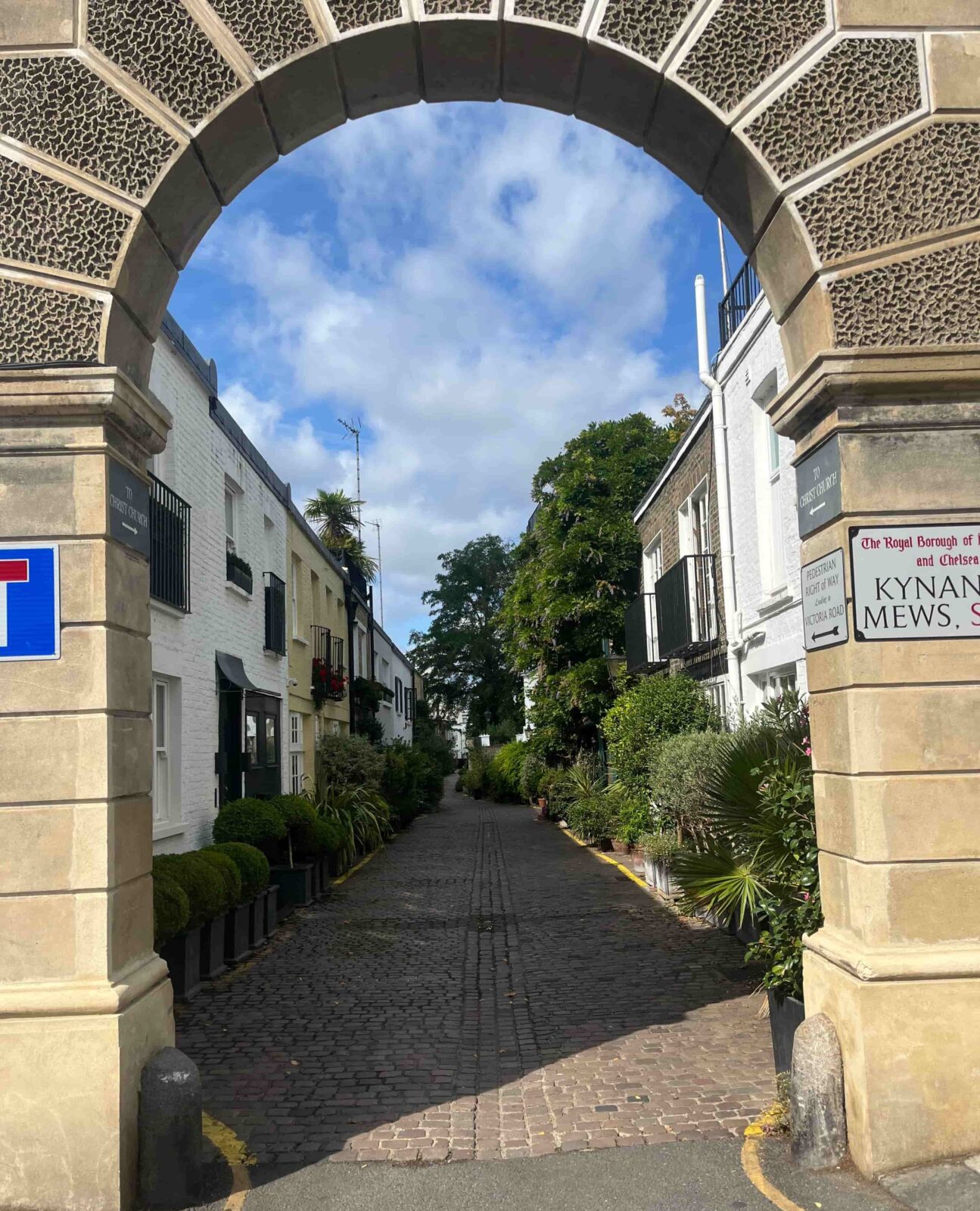 A shrubbery-lined, cobblestone street through an archway