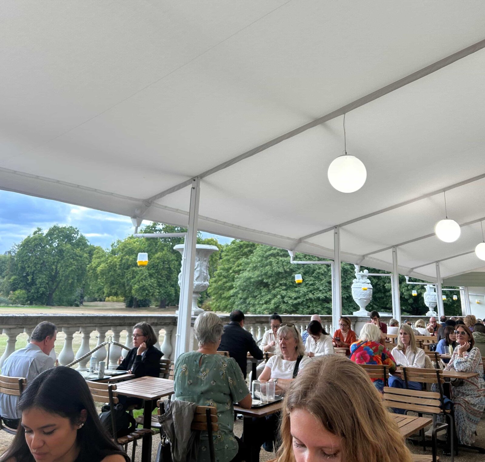 An outdoor cafe with people sitting at wooden benches under a white awning