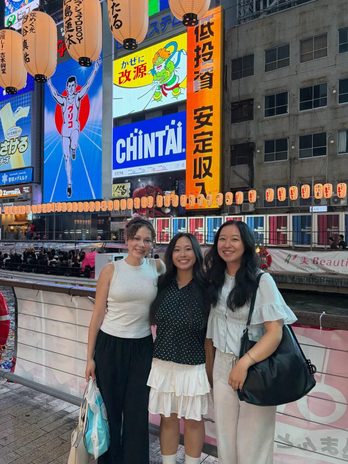 Three individuals leaning on a sidewalk railing, in a busy city, with bright signs and advertisements on the buildings behind them.