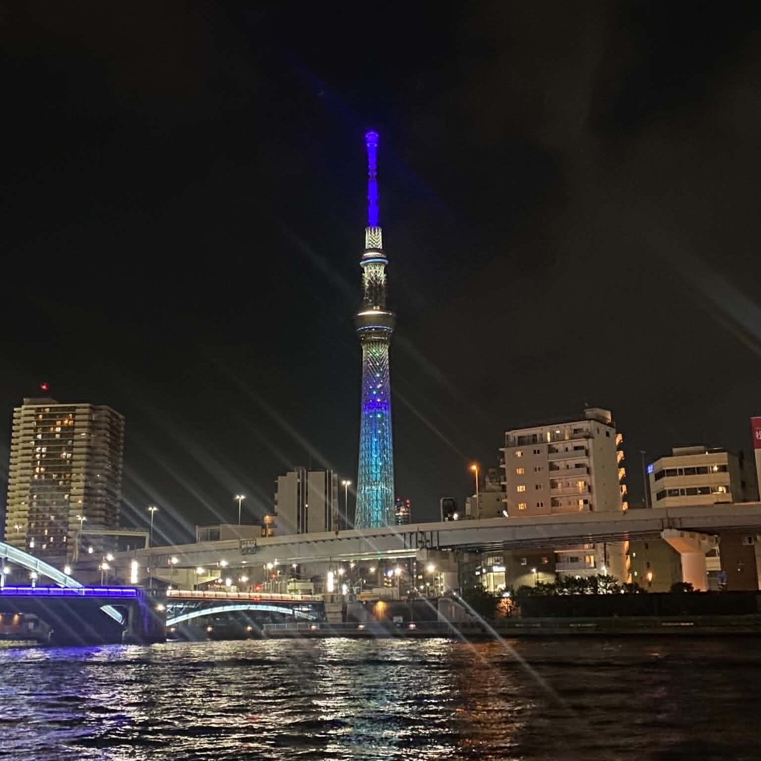 The tall Tokyo Skytree at night, lit blue and purple, viewed across water.