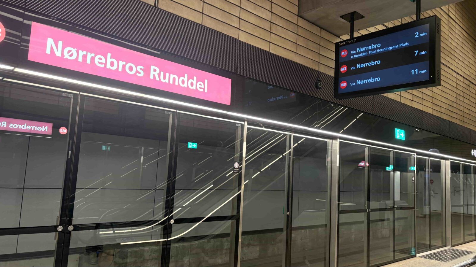 A clear platform screen door with a pink metro sign above it. There is also a TV displaying the upcoming trains.