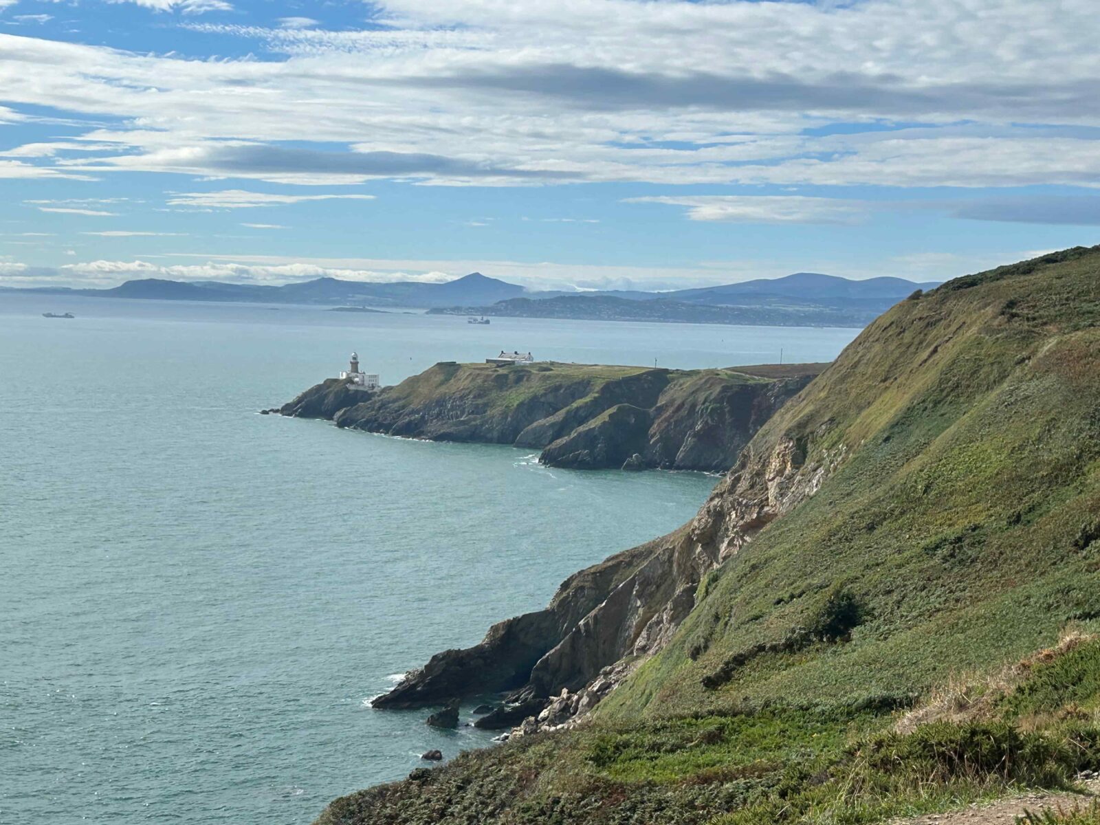 A rocky green cliffside with a lighthouse on a peninsula in the distance.