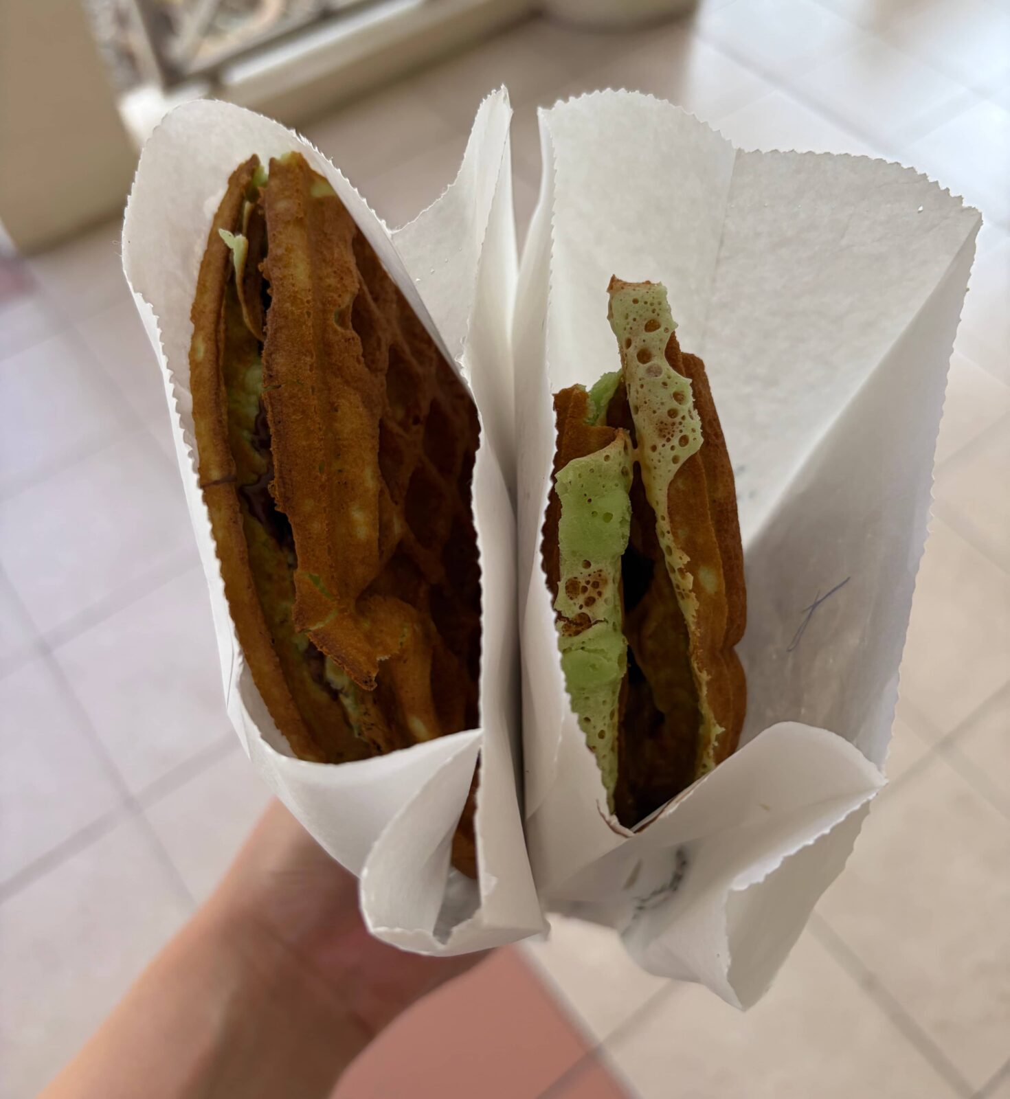 A hand holding two orders of pandan waffles, each in a white paper bag.