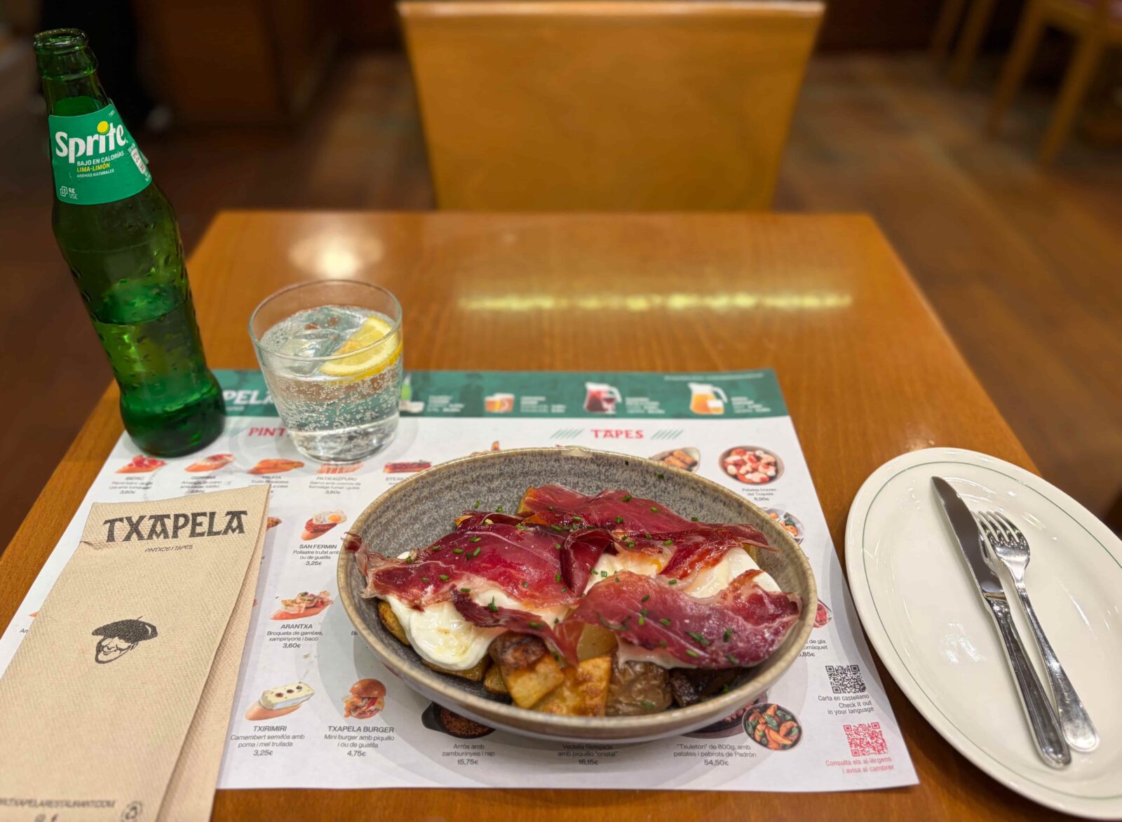 A small plate of tapas sits on top of a wooden table, paired with a glass bottle of Sprite